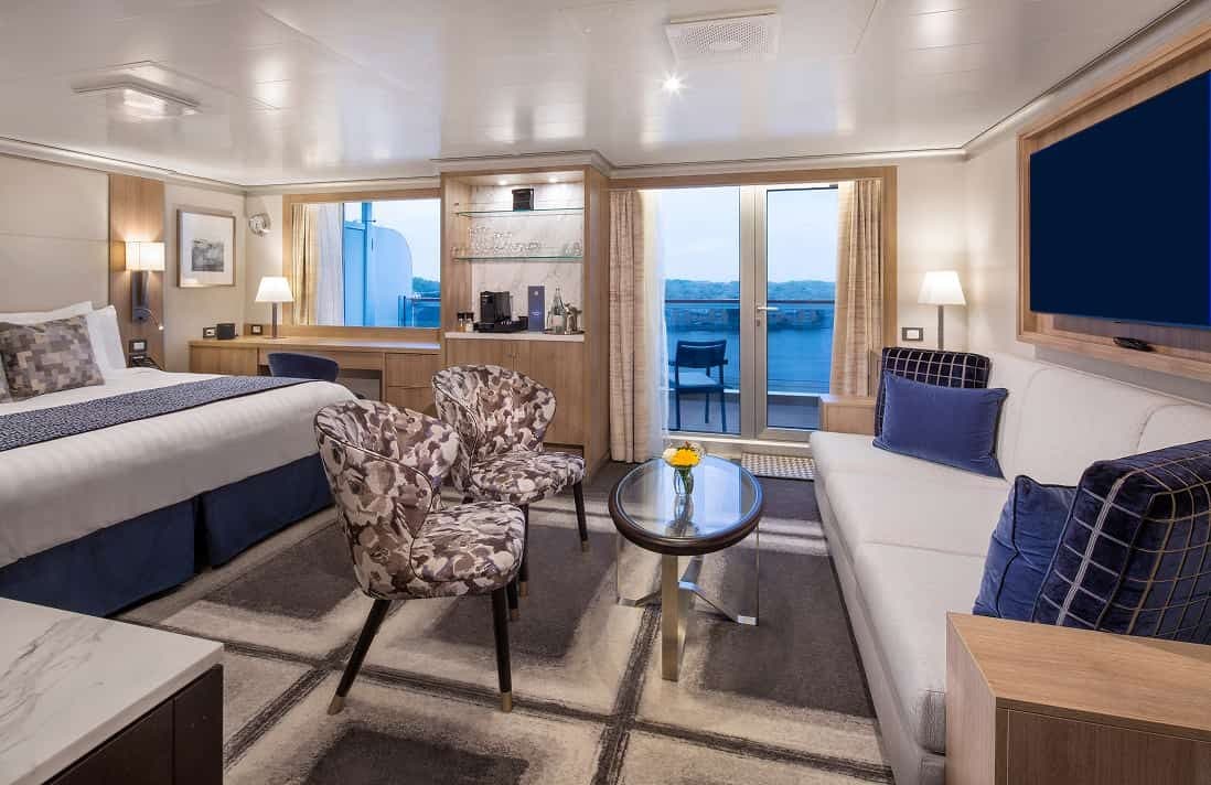 Neptune Suite Stateroom with king bed, patterned runner, two armchairs, sofa, coffee table, wall-mounted TV, and balcony on a Holland Americ