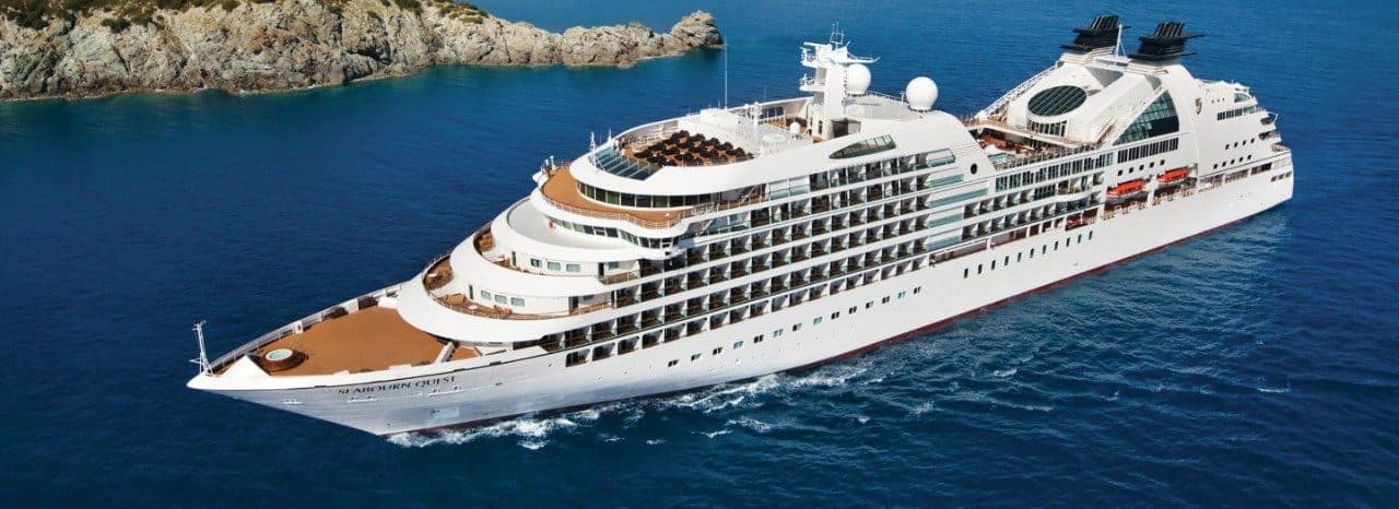 Seabourn Quest sailing along a rocky coastline in clear blue water, with multiple decks, sun loungers, and scenic island backdrop.